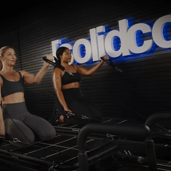 Two women exercising on reformer machines at Solidcore fitness studio with black walls